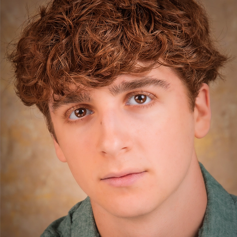 A young person with short, curly brown hair and fair skin looks directly at the camera with a neutral expression. They are wearing a green shirt, and the background is softly blurred in warm, earthy tones.