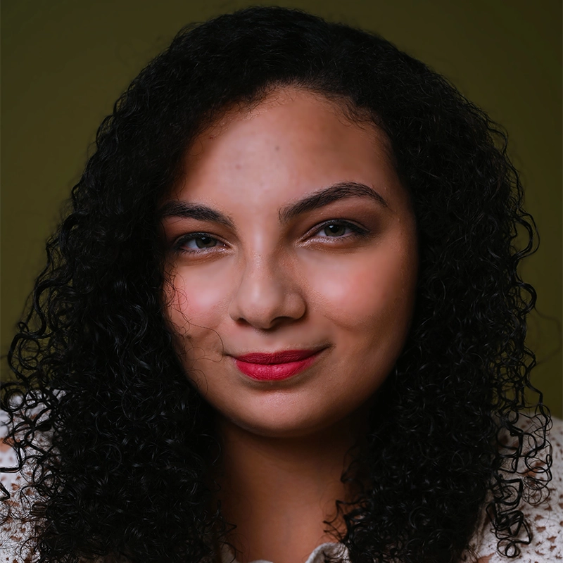 A woman with medium skin tone and curly black hair, resembling Farida Wahby, smiles slightly. She wears red lipstick, a cream-colored top, and has arched eyebrows. The plain olive green background and soft lighting highlight her features.