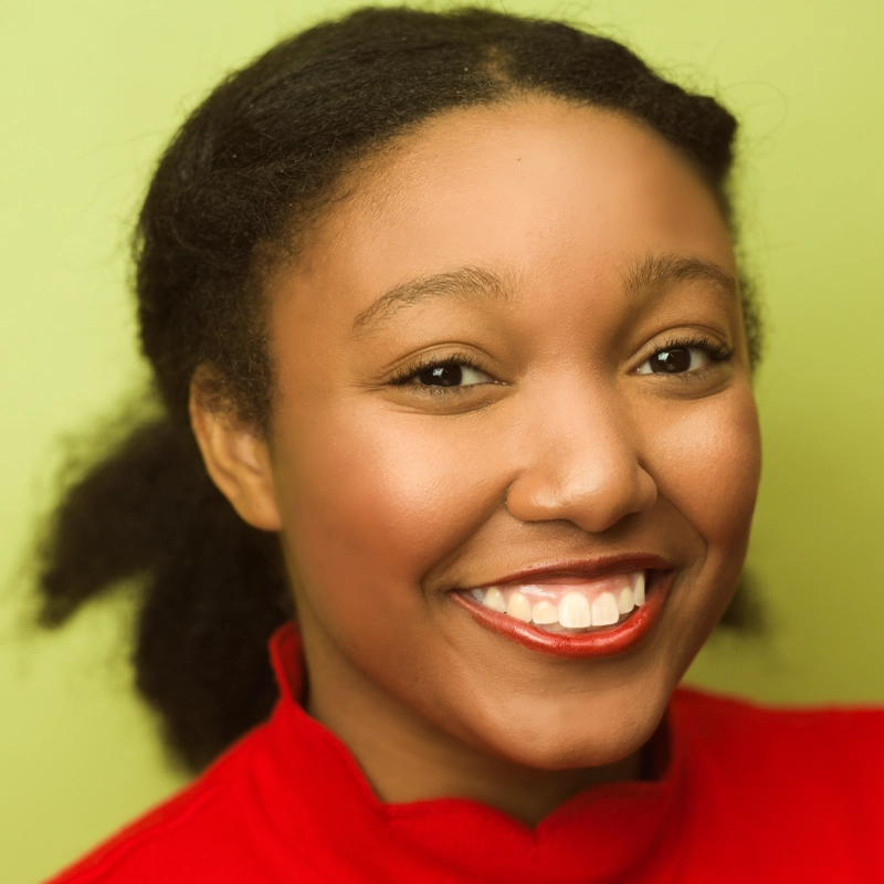 A woman with medium-dark skin and dark, textured hair pulled back smiles warmly. Ambroja Ferguson wears a bright red turtleneck and red lipstick. The light green background creates a cheerful and friendly portrait.