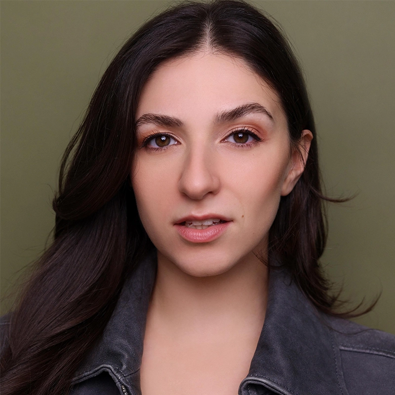 Gabriella Santalla, a woman with long brown hair and fair skin, looks directly at the camera. She has brown eyes, natural makeup, and slightly parted lips. She wears a dark gray jacket against an olive green background.
