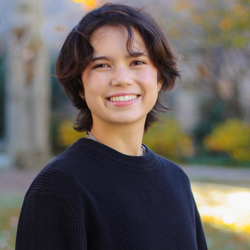 A person with short, wavy dark hair and light skin smiles outdoors. They wear a black, ribbed sweater and a chain necklace. The blurred background shows autumn trees with yellow leaves and soft sunlight.