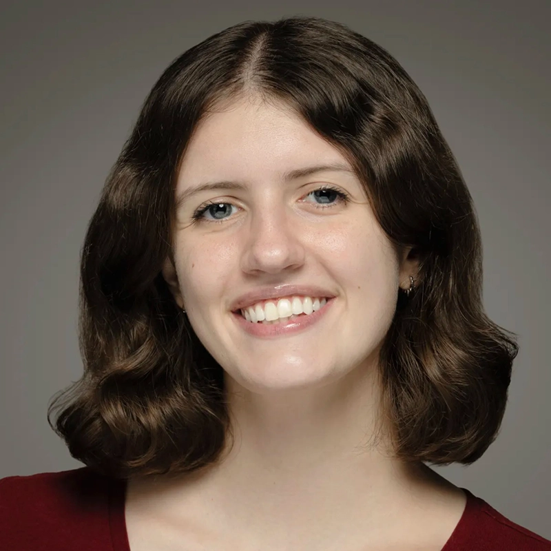 A young woman, Farida Wahby, with medium-length wavy brown hair smiles warmly at the camera. She has fair skin, blue eyes, and wears a burgundy top. The simple gray gradient background keeps the focus on her face.