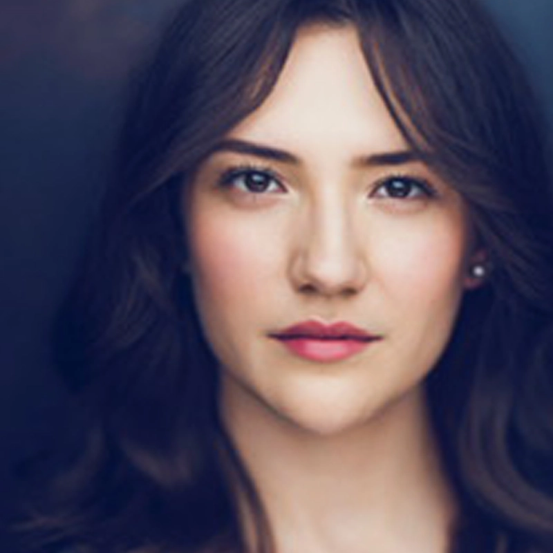 Karina Gallagher, a young woman with long, wavy brown hair and fair skin, looks directly at the camera. She has brown eyes, natural makeup, and pink lips. The softly blurred dark background highlights her calm, composed face.