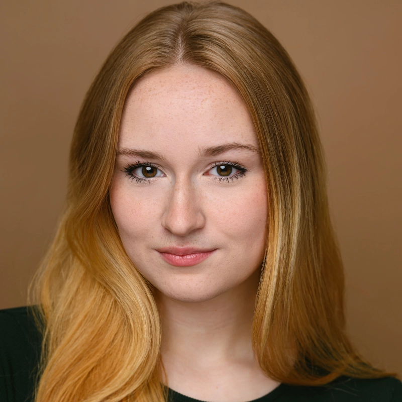 A young woman with long, straight blonde hair and fair skin is facing the camera. She has brown eyes, light makeup, and a slight smile. She wears a dark green top. The background is a solid, warm brown color.