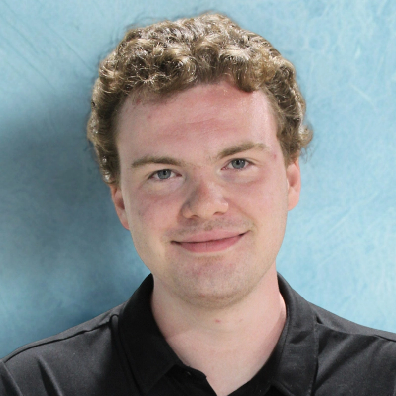 A young man with light skin and short, curly brown hair smiles at the camera. Gavin Matthias is wearing a black collared shirt and stands in front of a textured light blue background.