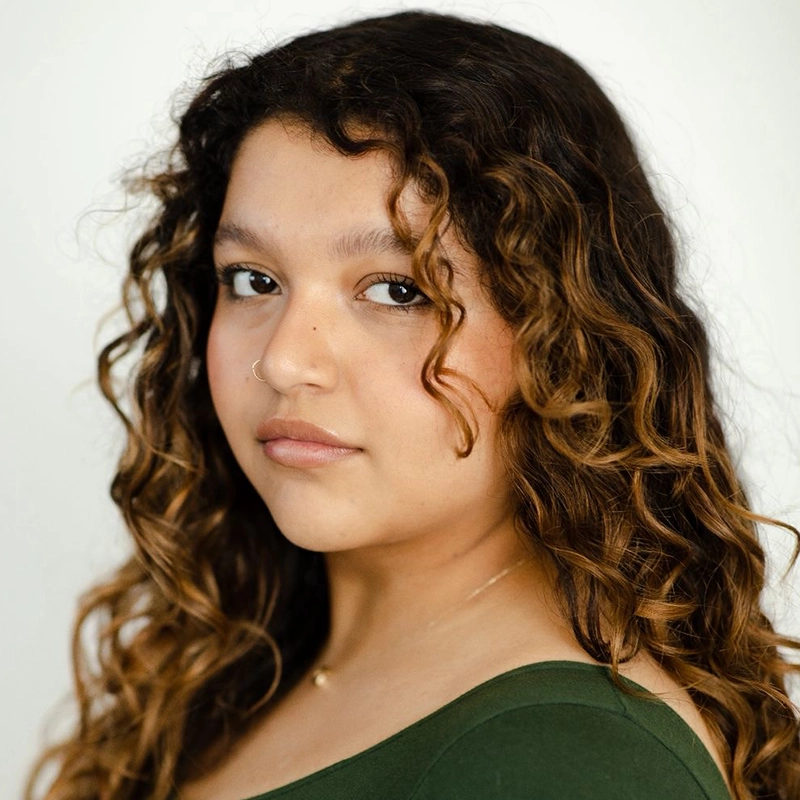A young person with long, curly brown hair and light skin looks slightly over their shoulder at the camera. Wearing a green top and a small nose ring, Genesis Aguila exudes calm confidence against a plain, light background.