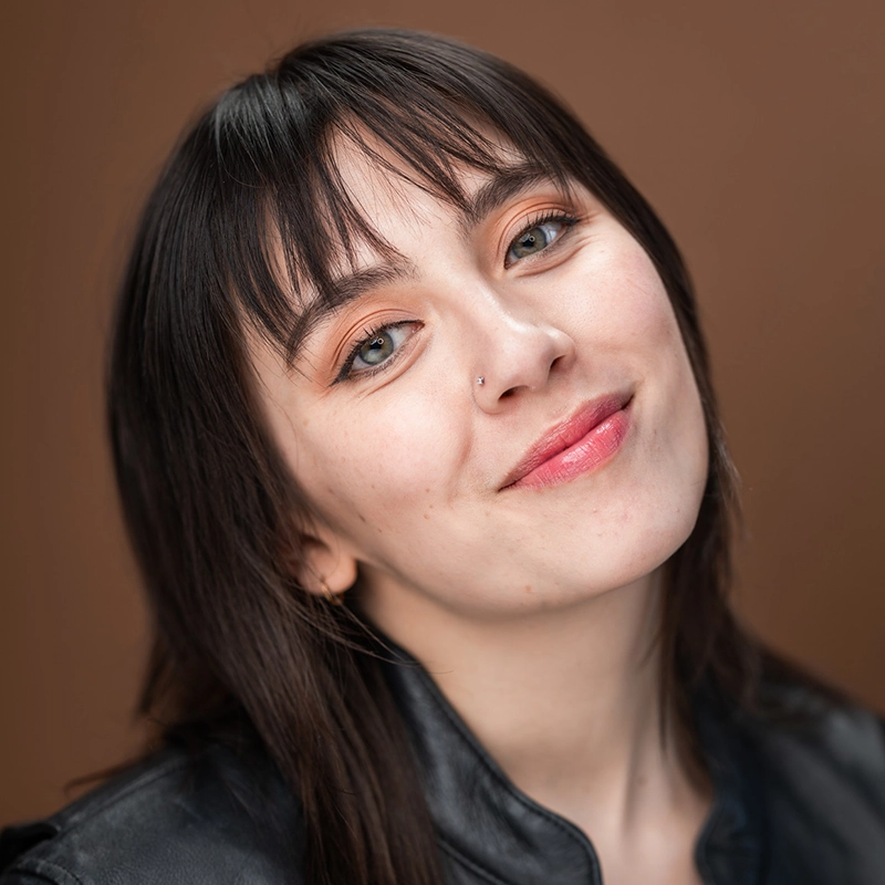 Genevieve Inez, a young woman with fair skin and straight dark brown hair, smiles softly. She has light makeup, a small nose piercing, and wears a black leather jacket against a plain brown background for a warm, neutral setting.