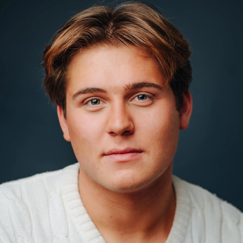 A young person with light skin, blue eyes, and short brown hair wears a white knit sweater. Alex Geoffroy has a neutral, relaxed expression and is posed against a dark blue background in this well-lit portrait that highlights their facial features.
