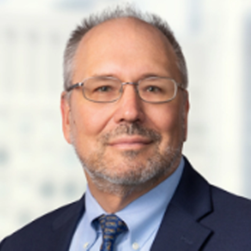 Kenneth Gerasimovich, a middle-aged man with short gray hair, glasses, and a trimmed beard, wears a navy blazer, light blue shirt, and blue patterned tie. He looks at the camera with a slight smile against a softly blurred light-toned background.