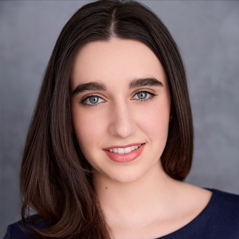 A young woman with long, straight brown hair and blue eyes smiles softly. She wears a navy blue top and is posed in front of a neutral gray background. Her makeup is natural, and her expression looks calm and friendly.