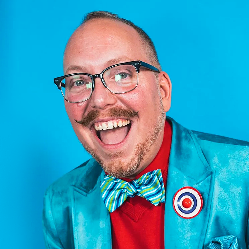 A smiling man with glasses, a mustache, and a short beard, wearing a shiny turquoise suit jacket, red shirt, and a colorful striped bow tie. He has a round pin on his lapel and stands in front of a bright blue background.