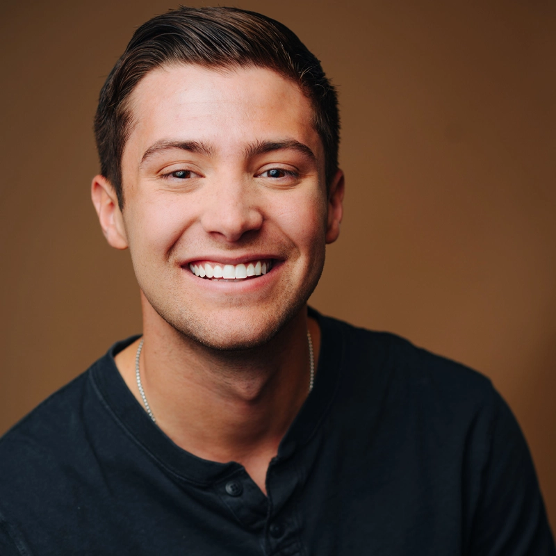 A young man with short brown hair smiles broadly, showing teeth. He wears a dark henley shirt and a thin necklace. The background is a solid, warm brown color, and the lighting highlights his face, giving a friendly and approachable appearance.