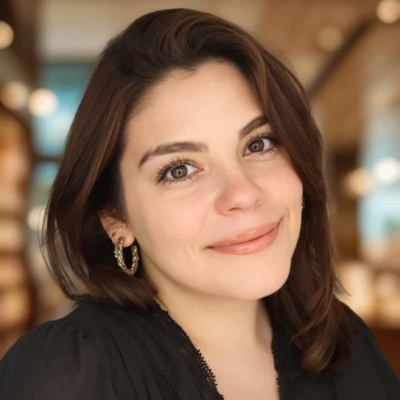 A young woman with light skin and shoulder-length brown hair smiles gently at the camera. Joanna Guarino wears gold hoop earrings and a black top. The background is softly blurred, suggesting an indoor setting with warm lighting.