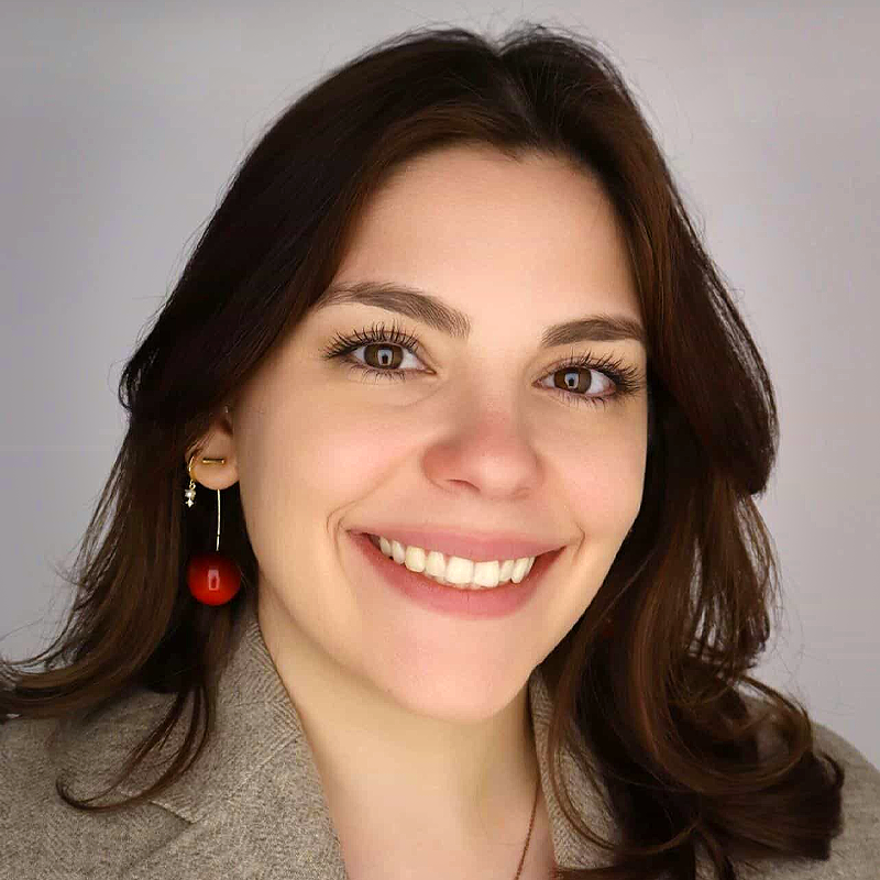 Joanna Guarino, with shoulder-length brown hair, smiles at the camera. She wears a tan blazer and red dangling earrings. The softly lit background draws attention to her warm expression and natural makeup.