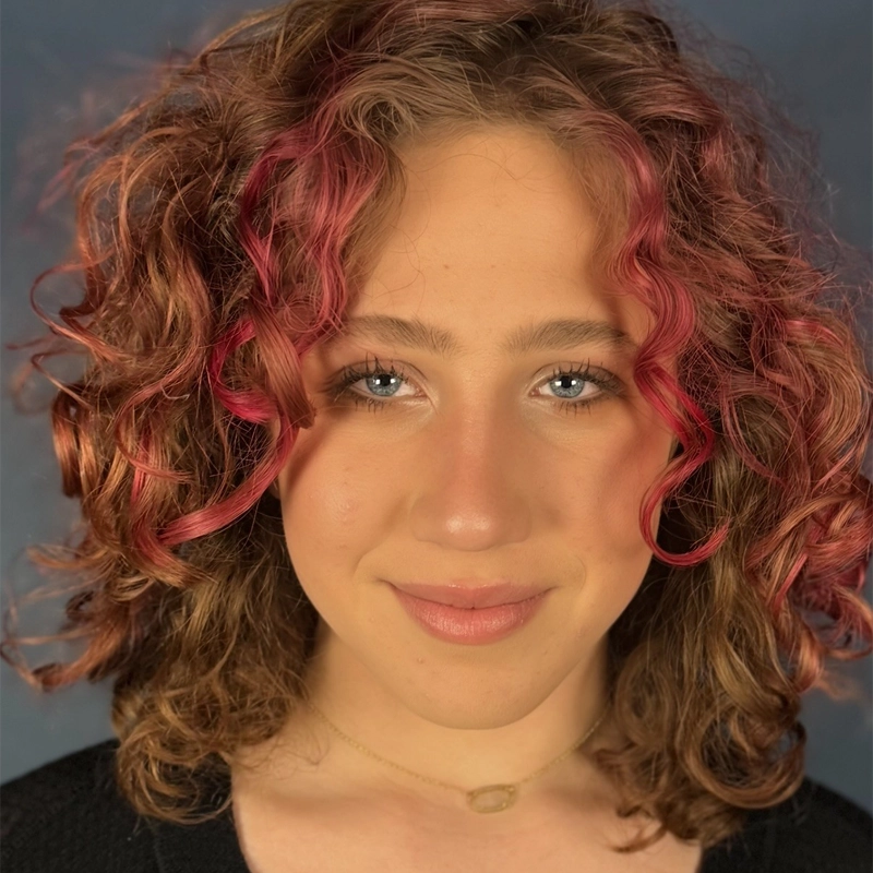 A person with light skin and curly, shoulder-length brown hair featuring pink highlights, smiling softly. They have blue eyes, natural makeup, and wear a gold necklace with an oval pendant. The background is softly blurred in blue tones.