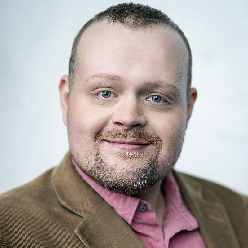 A smiling man with light skin, short brown hair, and a trimmed beard wears a tan corduroy blazer over a pink collared shirt. He faces the camera against a plain, light background.