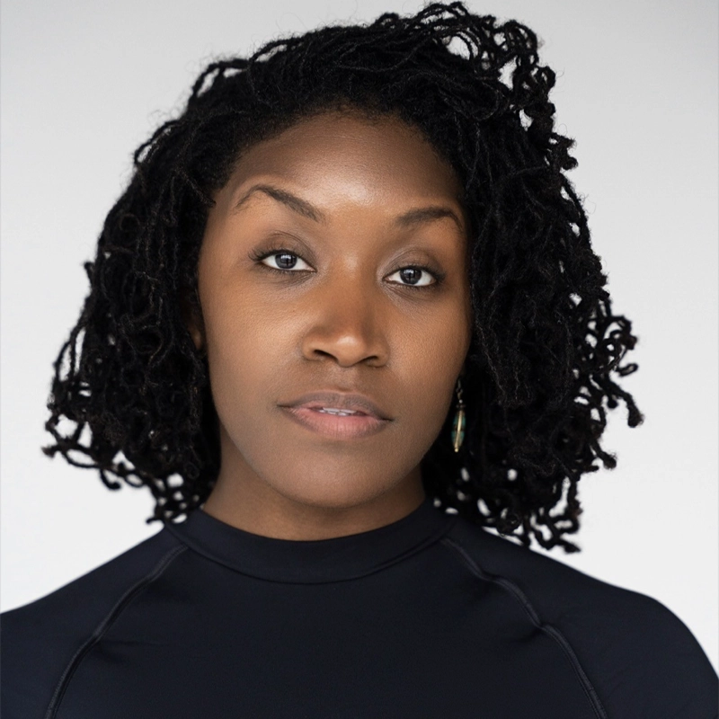 Kelsey Holley, a woman with medium brown skin and shoulder-length, curly black hair, looks directly at the camera with a neutral expression. She wears a black, high-neck top and small earrings against a plain, light-colored background.
