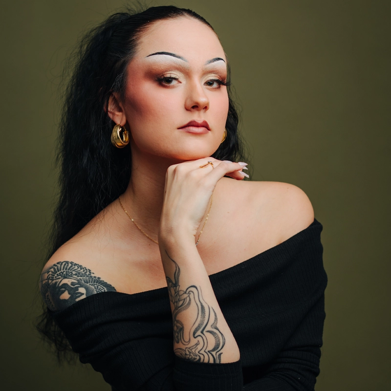 A woman with long dark hair and bold makeup poses with her chin resting on her hand. She wears gold hoop earrings, an off-shoulder black top, and has visible tattoos on her arm and shoulder. The background is plain olive green.