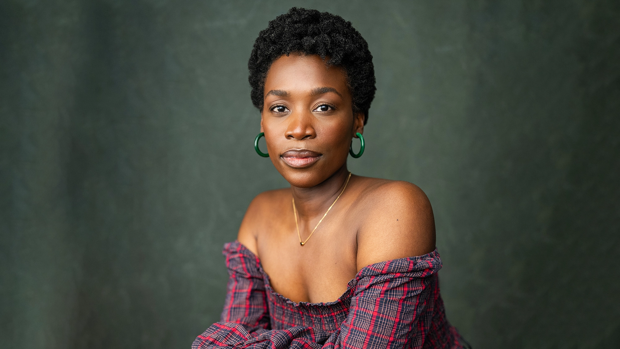 A woman with short curly hair and hoop earrings sits against a dark green background. Housso Sémon wears an off-the-shoulder, plaid, purple and red top and a delicate gold necklace, looking confidently at the camera with a neutral expression.