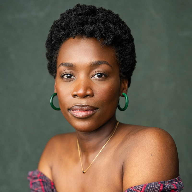 A woman with short, natural curly hair wears green hoop earrings, a gold necklace, and an off-the-shoulder plaid top. She looks confidently at the camera against a textured dark green background.