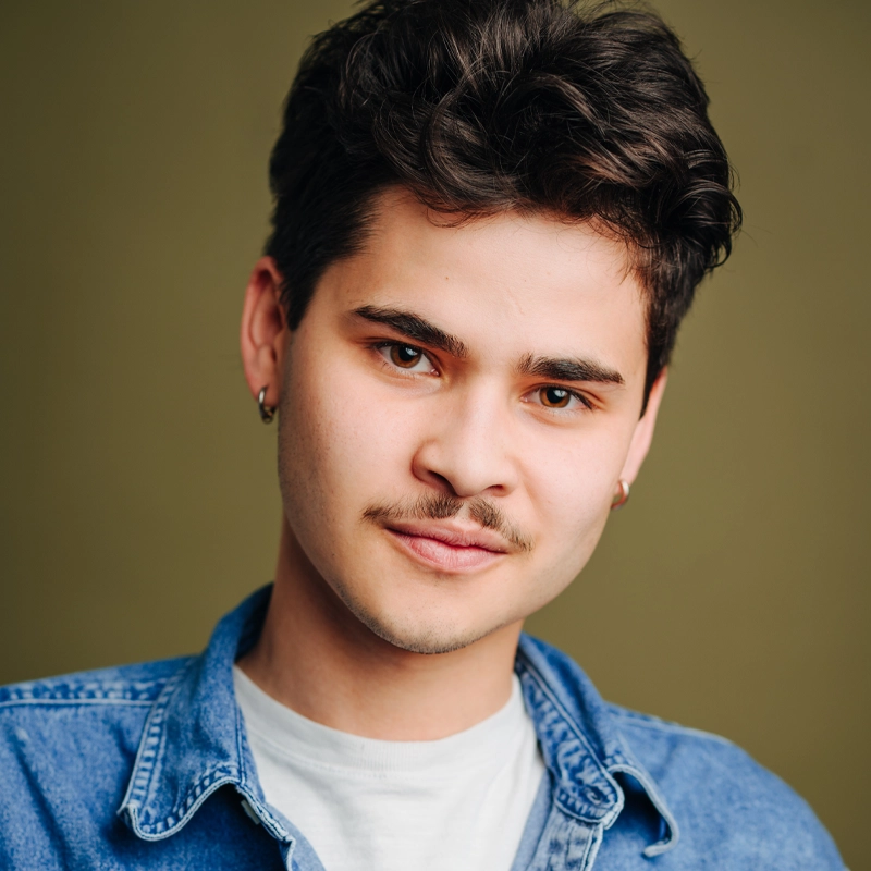 A young person with short, dark wavy hair and a faint mustache looks at the camera, wearing small hoop earrings, a denim shirt over a white t-shirt, and standing in front of an olive green background.
