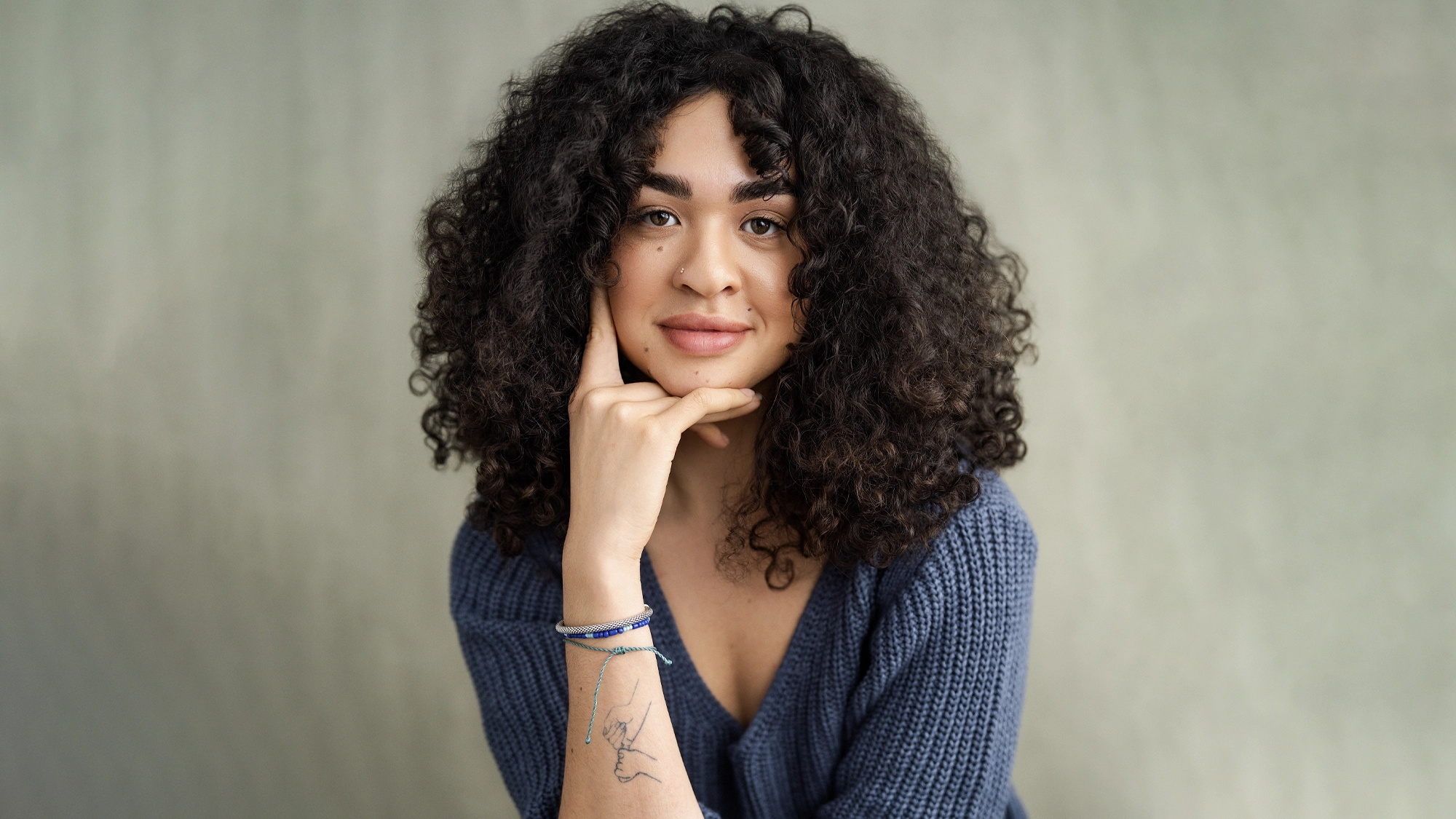 A person with curly dark hair and light skin, resembling Ilia Isorelýs Paulino, wears a textured blue sweater, resting their chin on one hand with a gentle smile. A line drawing tattoo is visible on their wrist against a subtle, neutral background.