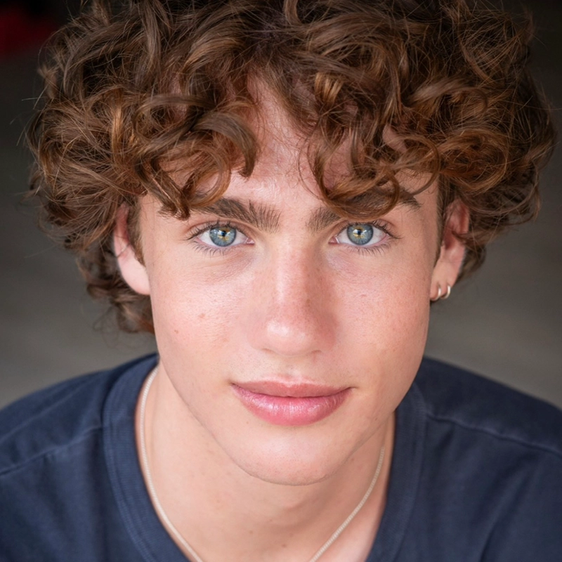 Jett Roberts, a young person with curly brown hair and blue eyes, looks directly at the camera, wearing a dark shirt, thin necklace, and small hoop earring in the left ear. The background is neutral and out of focus.