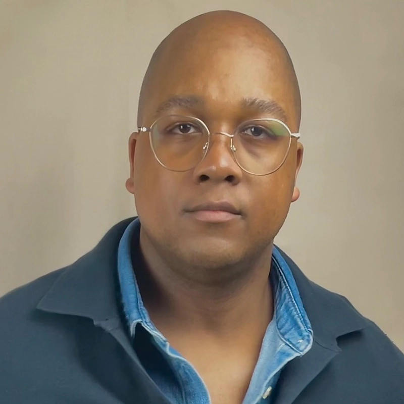 Jett Roberts, with a shaved head and glasses, looks straight at the camera with a neutral expression. They wear a light blue shirt under a dark jacket against a plain beige background.