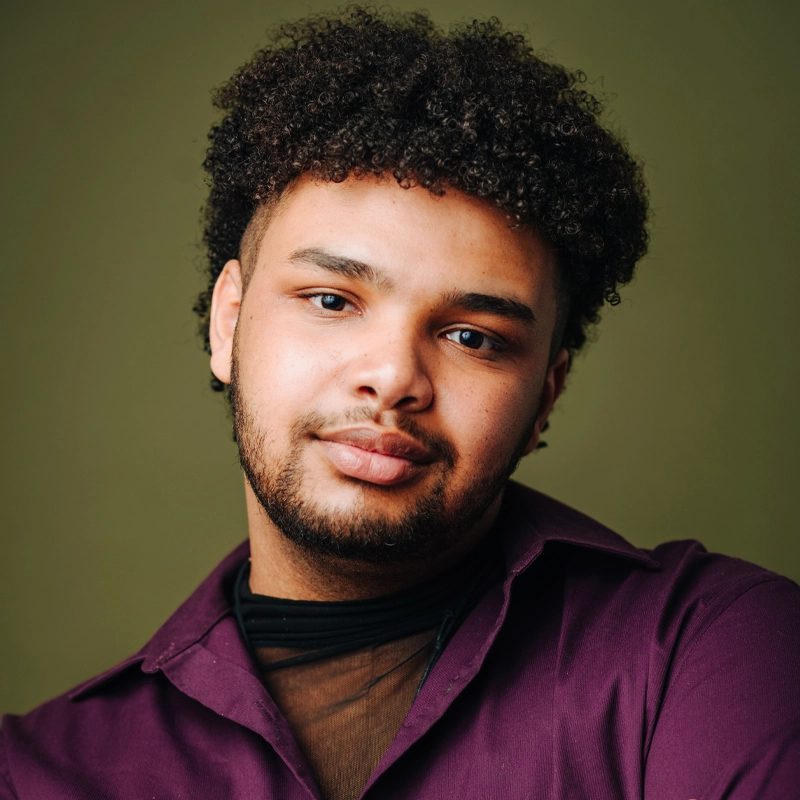 Malik Jallow, a young man with curly dark hair and a short beard, looks into the camera with a soft smile. He wears a sheer black top under a deep purple collared shirt against a plain olive green background, evenly lit to highlight his features.