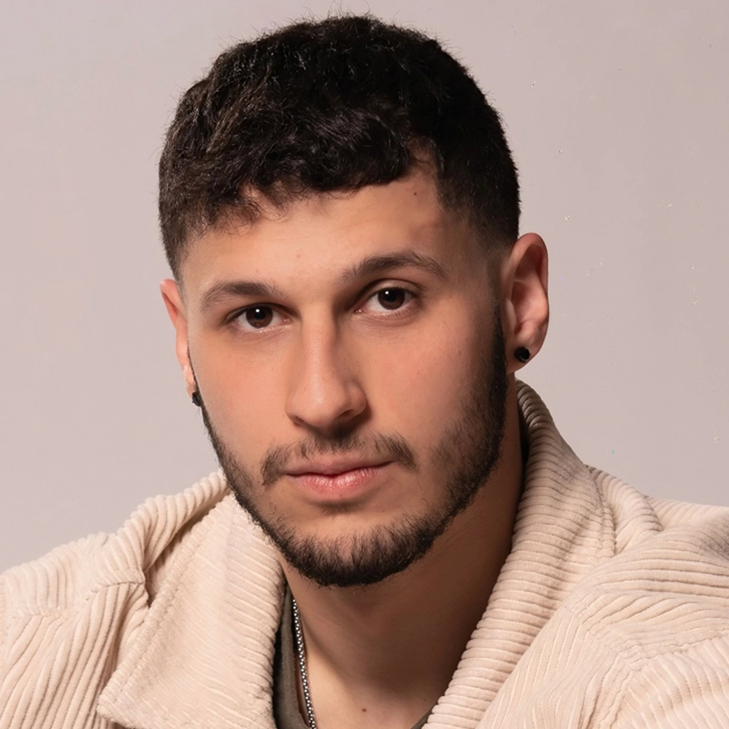 A young man with short dark hair, trimmed beard, and mustache looks directly at the camera. He wears a light beige corduroy jacket over a light shirt, a small black earring, and a silver necklace, with a neutral background.