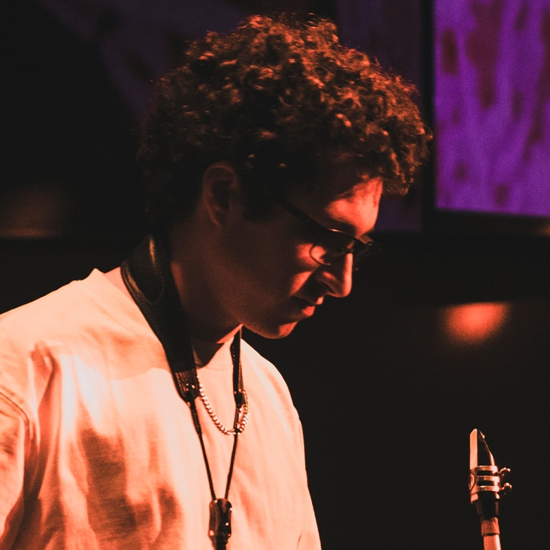 Annie Thomas, with curly hair and glasses, stands under warm, reddish stage lighting in a light shirt and necklace. She looks down thoughtfully, part of a musical instrument—possibly a clarinet or saxophone—visible in the foreground.
