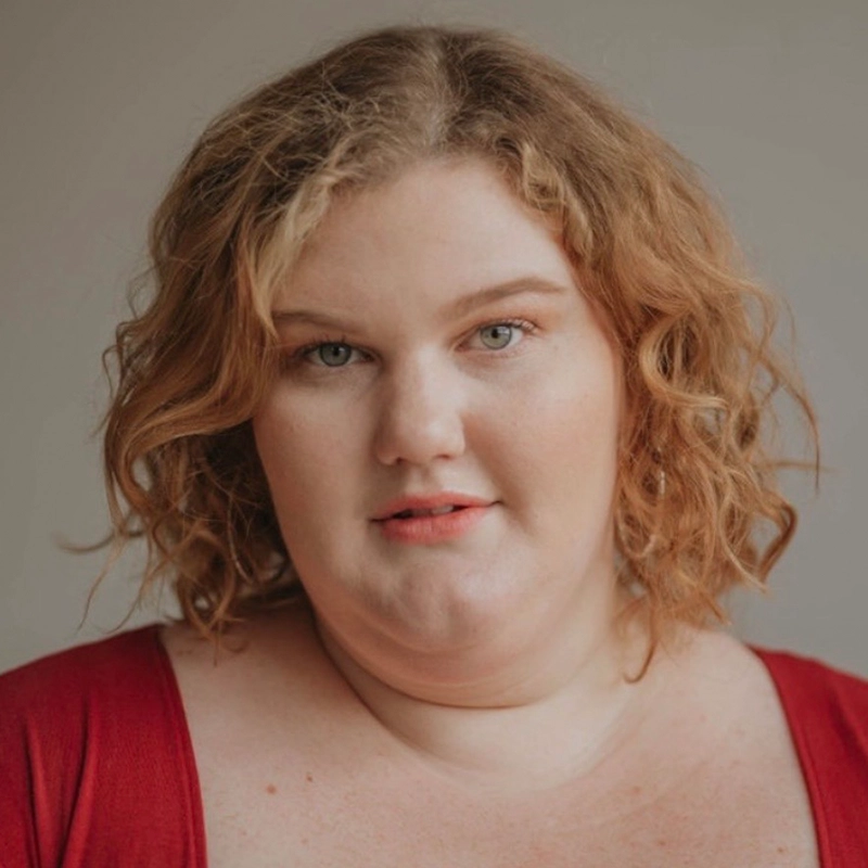 A woman with wavy, shoulder-length blonde hair and fair skin looks at the camera with a neutral expression. Jett Roberts wears a deep red top and natural makeup against a plain, softly lit background.