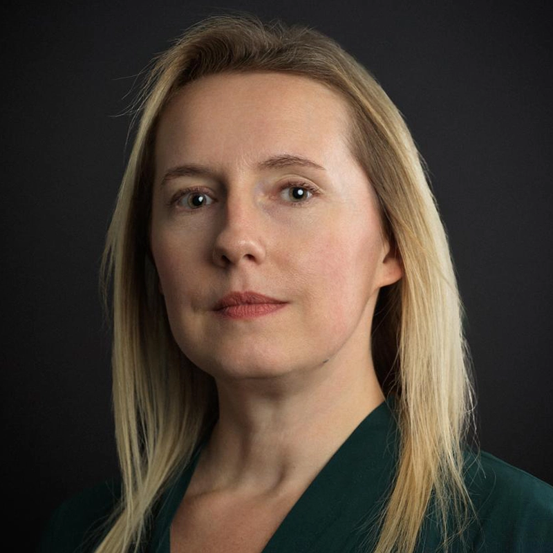 A woman with light skin and straight blonde hair, wearing a dark green top, looks directly at the camera with a neutral expression against a plain dark background. The soft lighting highlights her facial features in this Xavier Wilson-inspired portrait.