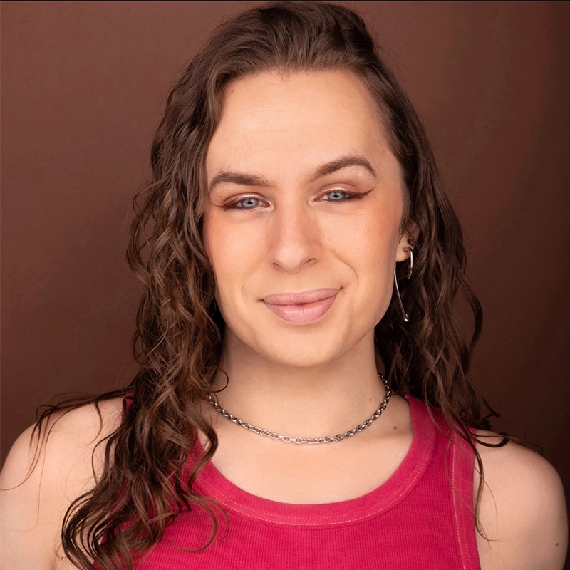 Jett Roberts, with long, wavy brown hair and light skin, smiles warmly at the camera. They wear a sleeveless red top, a silver chain necklace, and a small earring against a solid brown background.