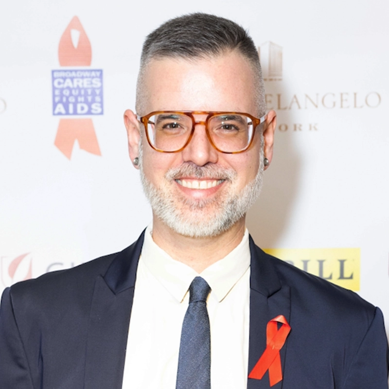 A smiling person with short gray hair, glasses, and a beard wears a dark suit, white shirt, and dark tie. A red awareness ribbon is pinned to their lapel. The background features various logos, including Broadway Cares and Brooke Cassatto graphics.