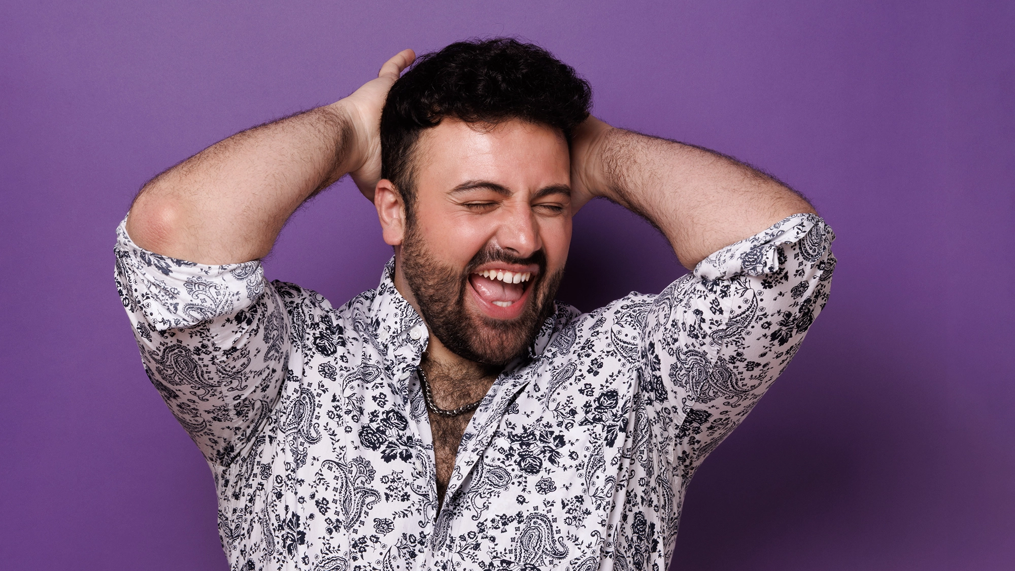 A bearded man wearing a patterned white and black shirt stands in front of a purple background, smiling broadly with his eyes closed and hands placed behind his head in a joyful, relaxed pose, channeling Pop Diva vibes like Joe Marano.