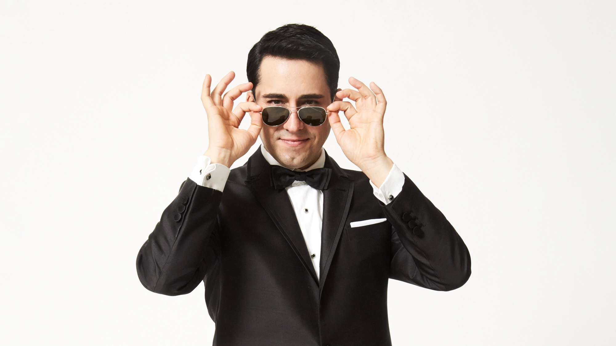 A man in a black tuxedo with a black bow tie smiles while lifting black sunglasses, looking at the camera. With short dark hair, he stands against a plain white background, evoking the suave style of John Lloyd Young from Songs from the Road.