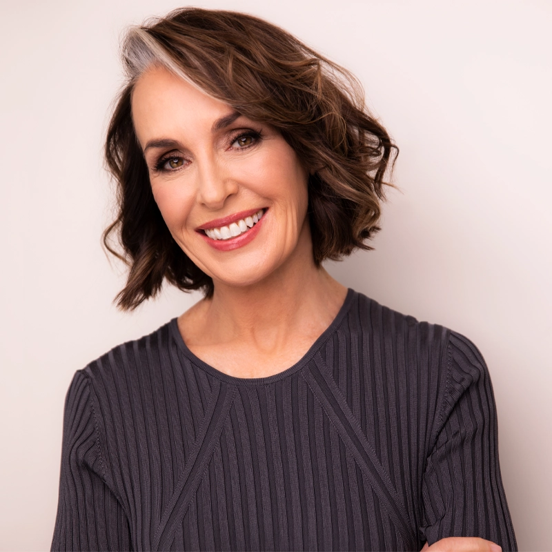 A smiling middle-aged woman, Maree Johnson, with short, wavy brown hair featuring a streak of gray. She is wearing a ribbed, dark gray top and stands in front of a plain light background, looking directly at the camera with a friendly expression.
