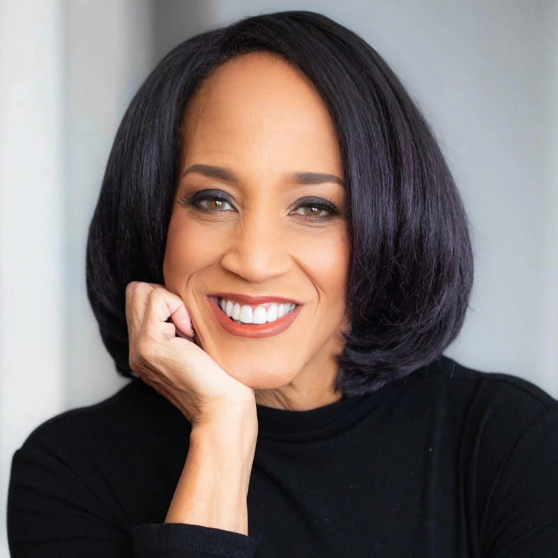 Michelle Johnson, a middle-aged woman with straight, shoulder-length dark hair, smiles warmly at the camera. She rests her chin on her hand and wears a black top. The softly blurred background in neutral tones keeps the focus on her face.