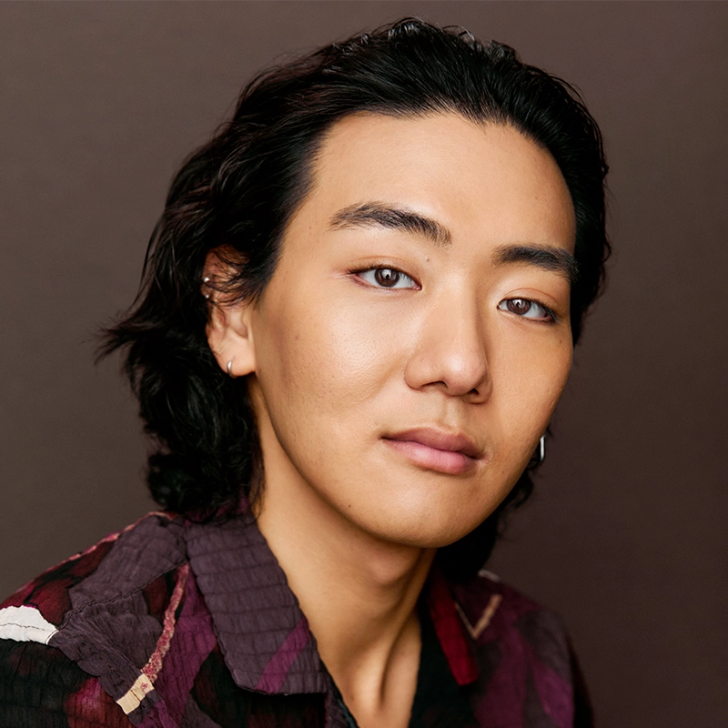 Gabriella Santalla, a young person with medium-length, wavy dark hair and fair skin, gazes softly at the camera. They wear a maroon and black patterned shirt and a small hoop earring, set against a plain dark brown background.