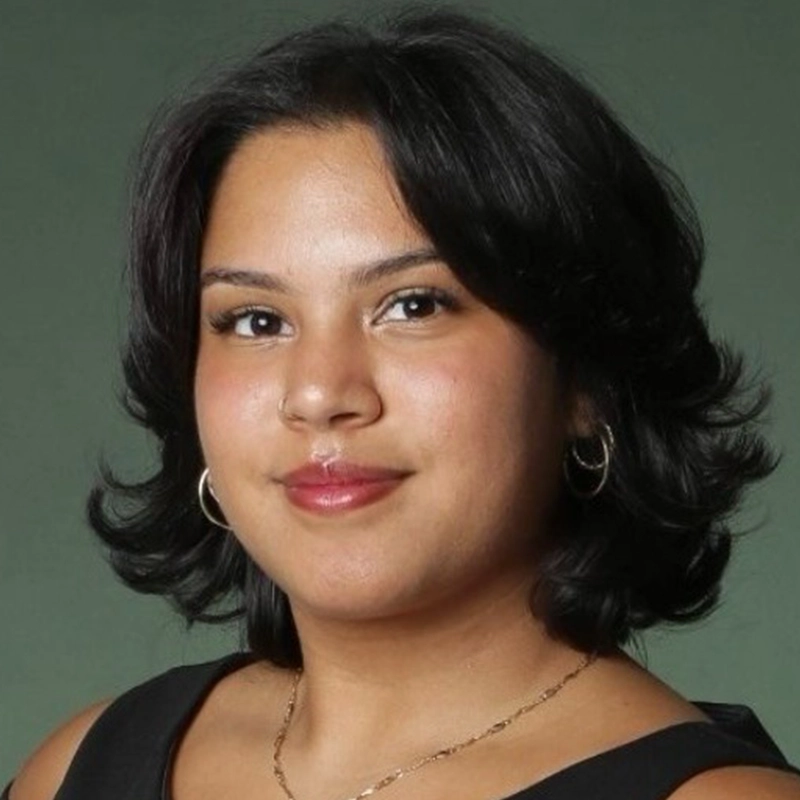 A young woman with medium skin tone and short, wavy black hair smiles gently at the camera. She wears gold hoop earrings, a thin gold necklace, and a sleeveless black top by Danté X Johnson. The background is a solid muted green.