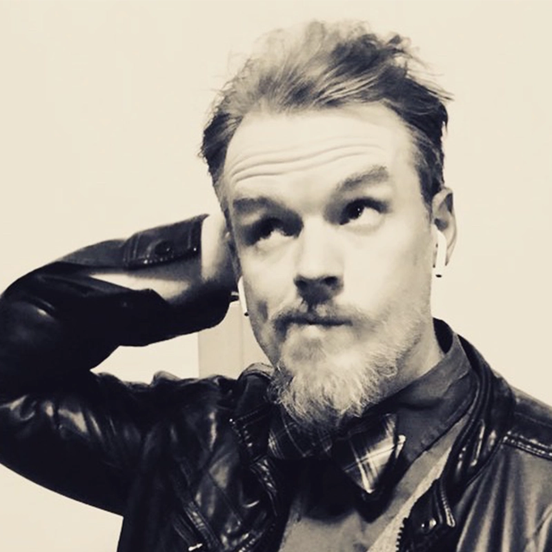 A man with light skin, a short beard, and mustache looks upward thoughtfully. Justin Fischer wears a leather jacket, plaid shirt, and bowtie, with earbuds in and one hand behind his head in this black and white portrait.