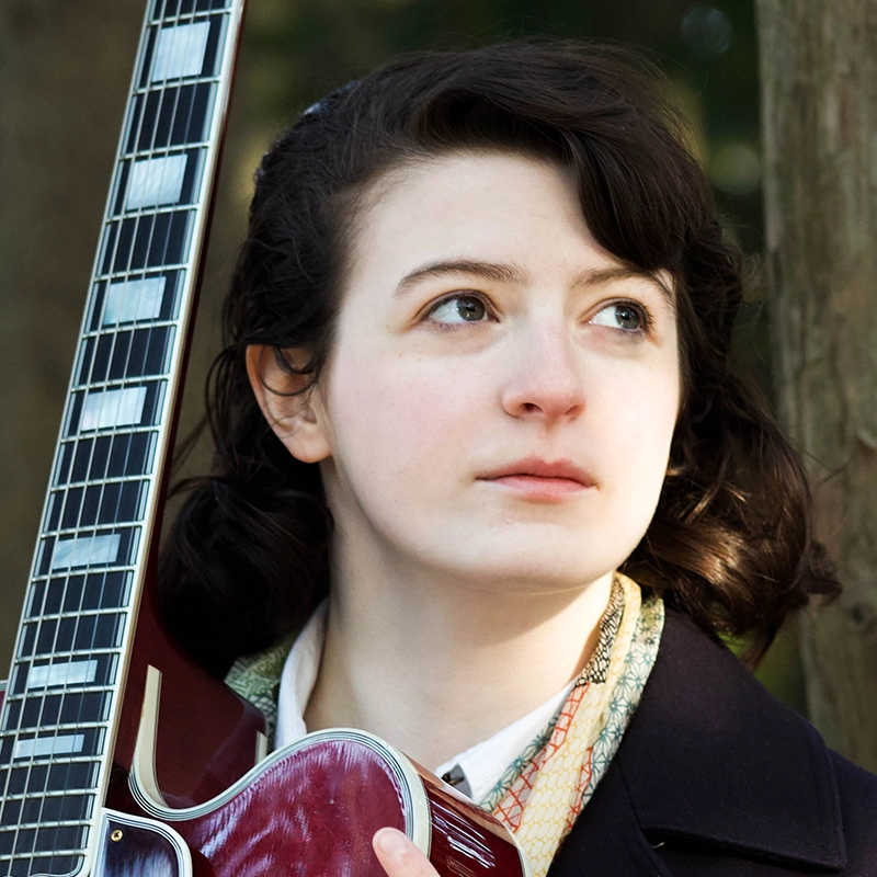 Genevieve Inez, a young woman with short dark hair, holds a guitar vertically in front of her. She gazes thoughtfully to the side, dressed in a dark coat and patterned scarf. The outdoor background features blurred trees and natural light.