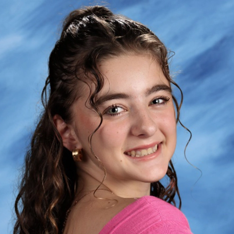 A young woman with long, wavy brown hair and blue eyes smiles at the camera. Farida Wahby wears a pink top and gold hoop earrings. The background is a blue, cloud-like studio backdrop, commonly used in school portraits.