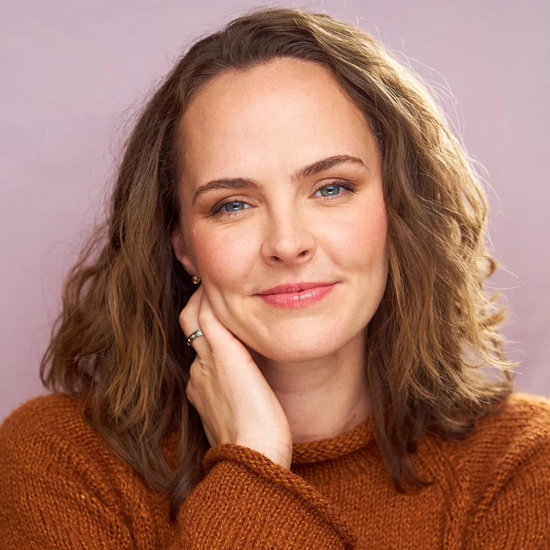 A woman with wavy, shoulder-length brown hair and blue eyes smiles gently. She wears a rust-colored sweater and rests her chin on her hand, showing a ring on her finger. The background is a soft pinkish-purple color.