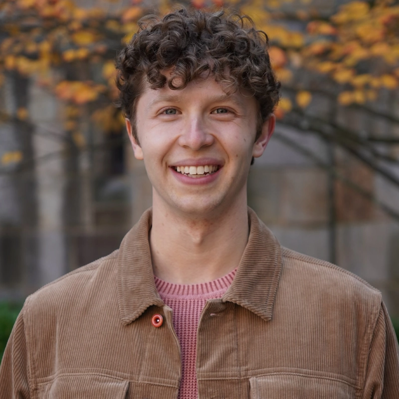 Joseph Kayne, a young man with short curly brown hair, smiles at the camera in a brown corduroy jacket over a pink sweater. Blurred autumn trees with orange leaves and a stone building create a warm, cheerful scene in the background.