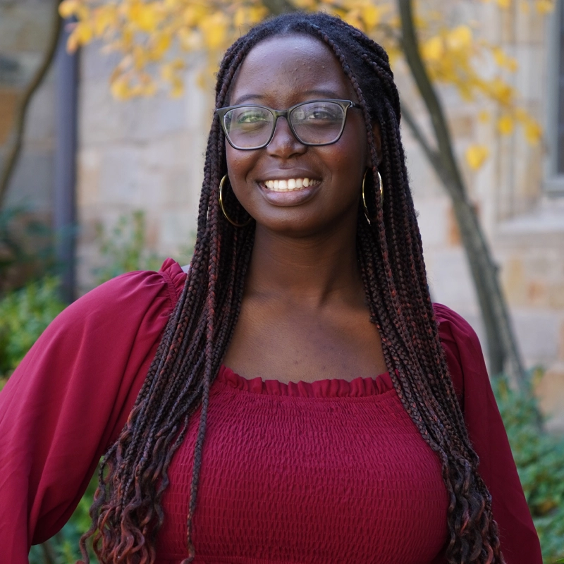 A woman with long braids and glasses, wearing large hoop earrings and a maroon ruched blouse, smiles outdoors in front of a stone wall and trees with yellow autumn leaves.