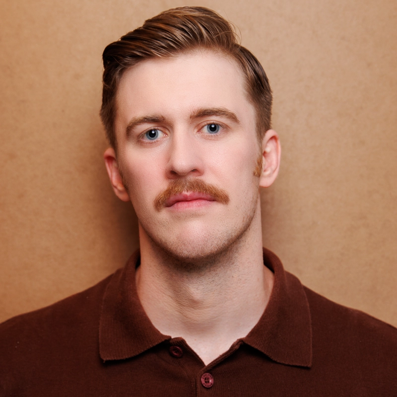 A man with light skin, short brown hair styled to the side, and a thick mustache—resembling Kieran Kelly—looks directly at the camera. He wears a brown collared shirt and stands before a plain tan background, his expression neutral.