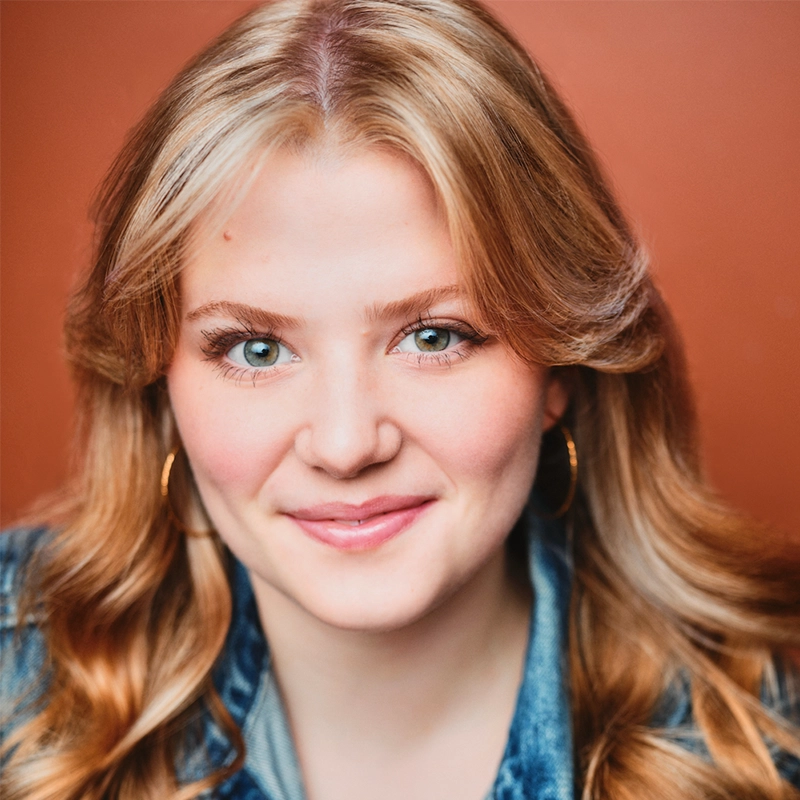 Farida Wahby, a young woman with wavy blonde hair and blue eyes, smiles gently at the camera. She wears gold hoop earrings and a denim jacket against a warm, reddish-brown background, giving her a friendly and approachable appearance.
