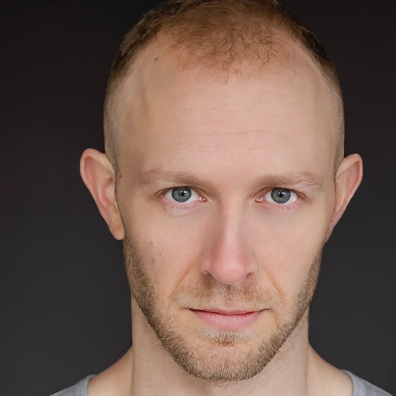 A man with short, light brown hair and a receding hairline stares directly ahead with a neutral expression. He has light skin, blue eyes, and a trimmed beard. He wears a grey shirt, and the background is solid black.
